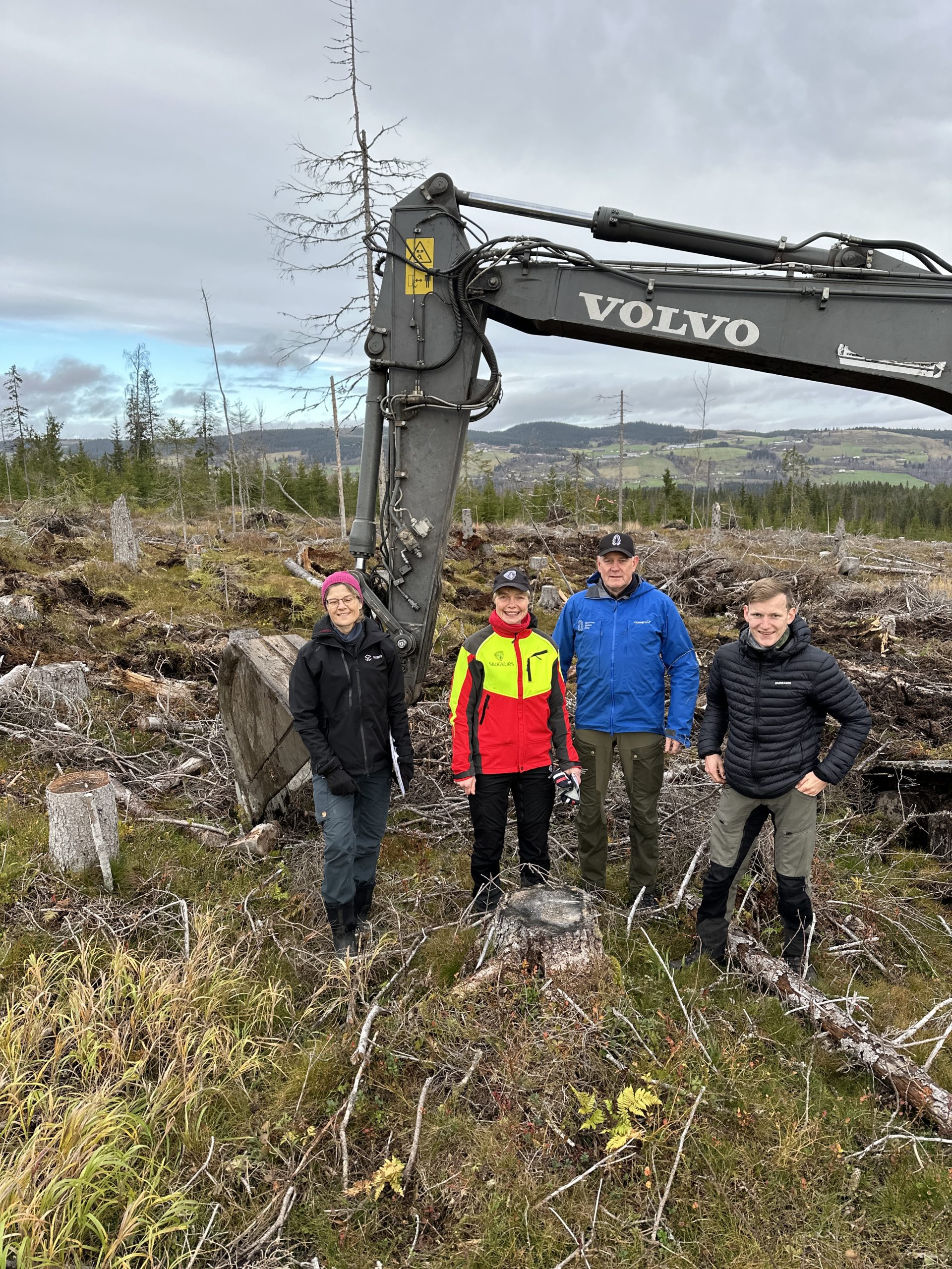 Gruppe personer foran gravemaskin på hogstflate.