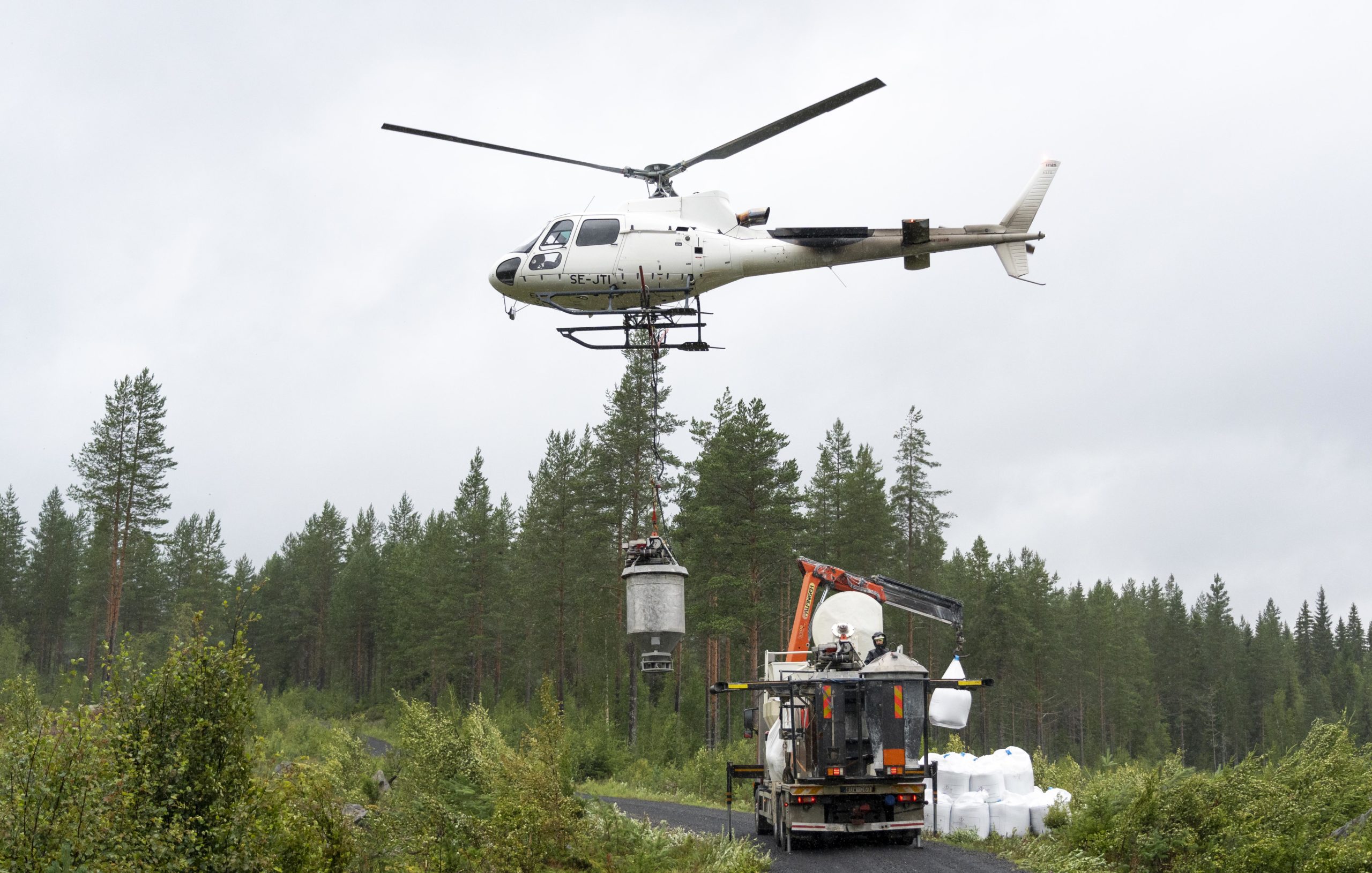 Helikopter med gjødseltank flyr lavt over lastebil med gjødsel.